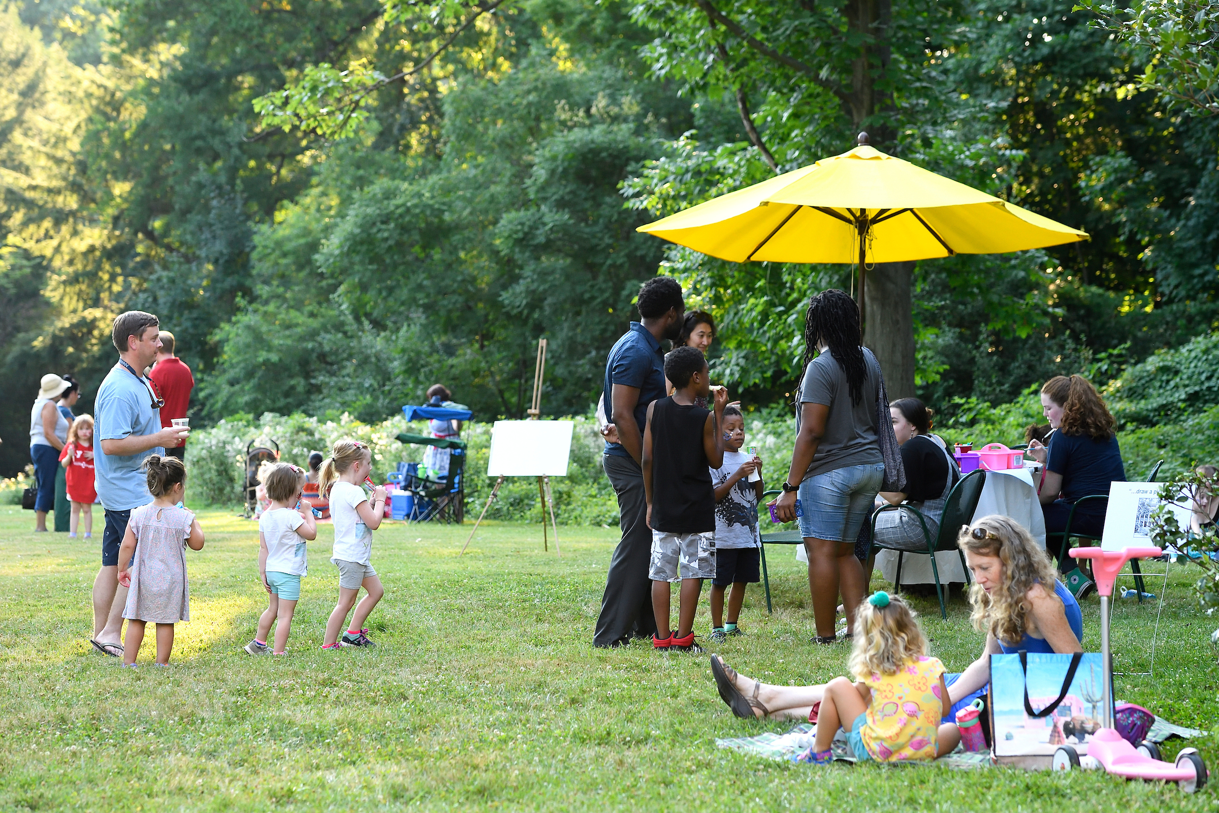 Clusters of children and adults congregate on Evergreen's front lawn. To the left, a table with chairs and a yellow umbrella is set up.