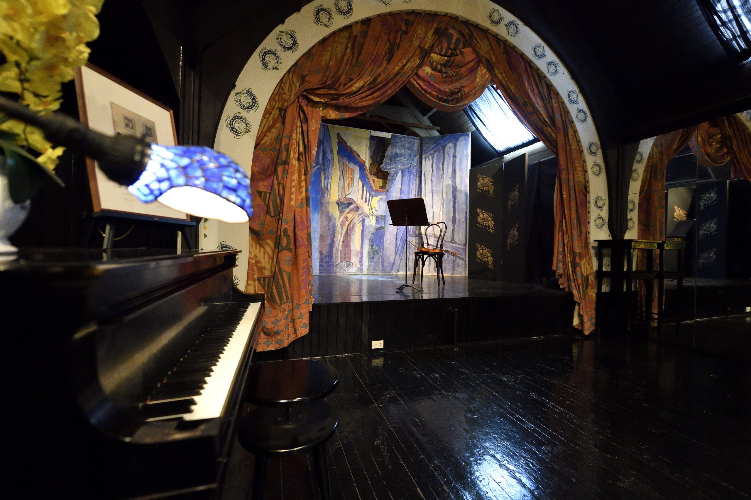 Bakst Theatre with a close up of baby grand piano keys. In the background, a single chair and music stand are on the stage. A violin rests in the chair's seat.