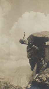 A sepia-toned image of a man standing on a piece of rock jutting out from a sheer cliff. Below him, a western valley yawns in the distance. Billowing cumulus clouds tower above and behind him.