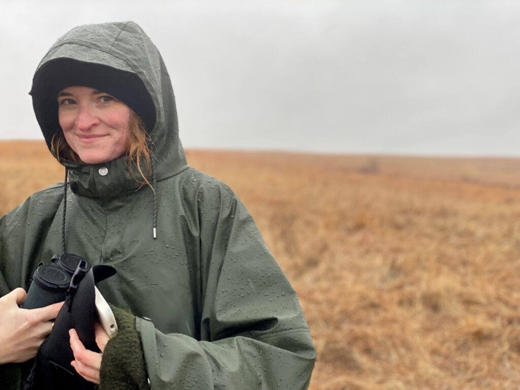 Sarah Kaizar, standing in a field, wearing rain gear, and holding a pair of binoculars.